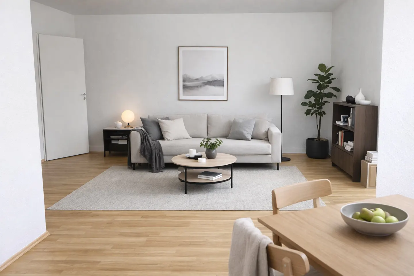 Bright living room with a beige sofa, round coffee table, and wooden dining table with a bowl of green apples.