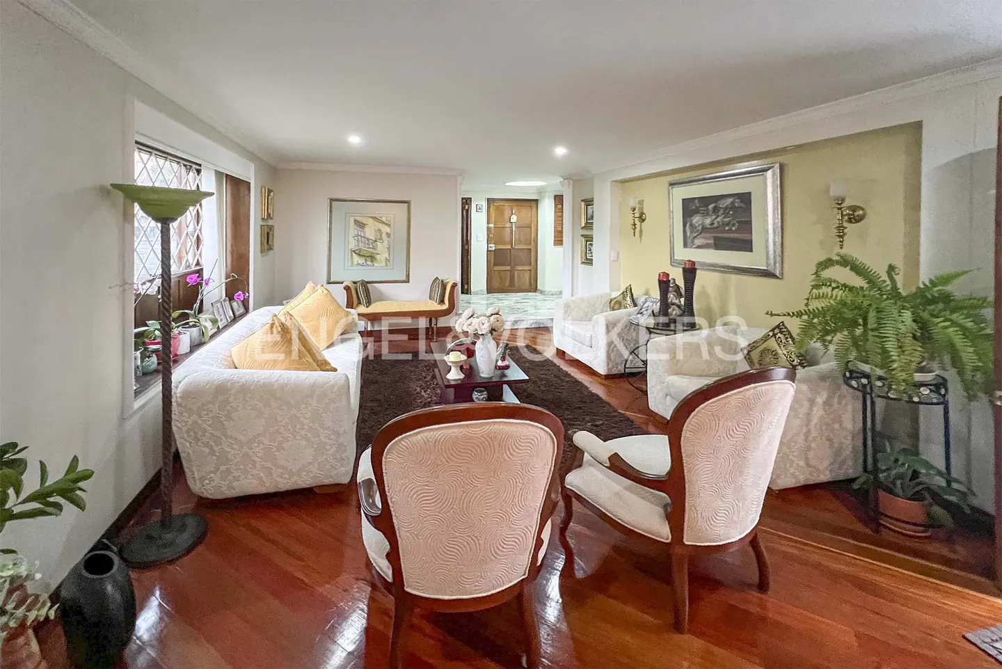 A living room with hardwood floors, white sofas, and two chairs facing away from the camera. A brown rug is in the center of the room.