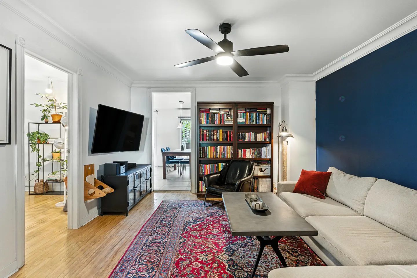 A living room with a blue accent wall, a beige sofa, a dark wood bookcase, and a red patterned rug. A black ceiling fan is overhead.