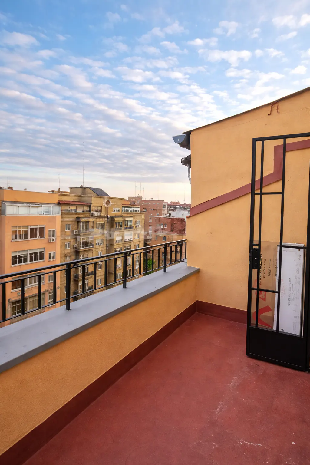 Rooftop terrace with red flooring, black railing, and open black metal gate. City buildings and cloudy sky in the background.
