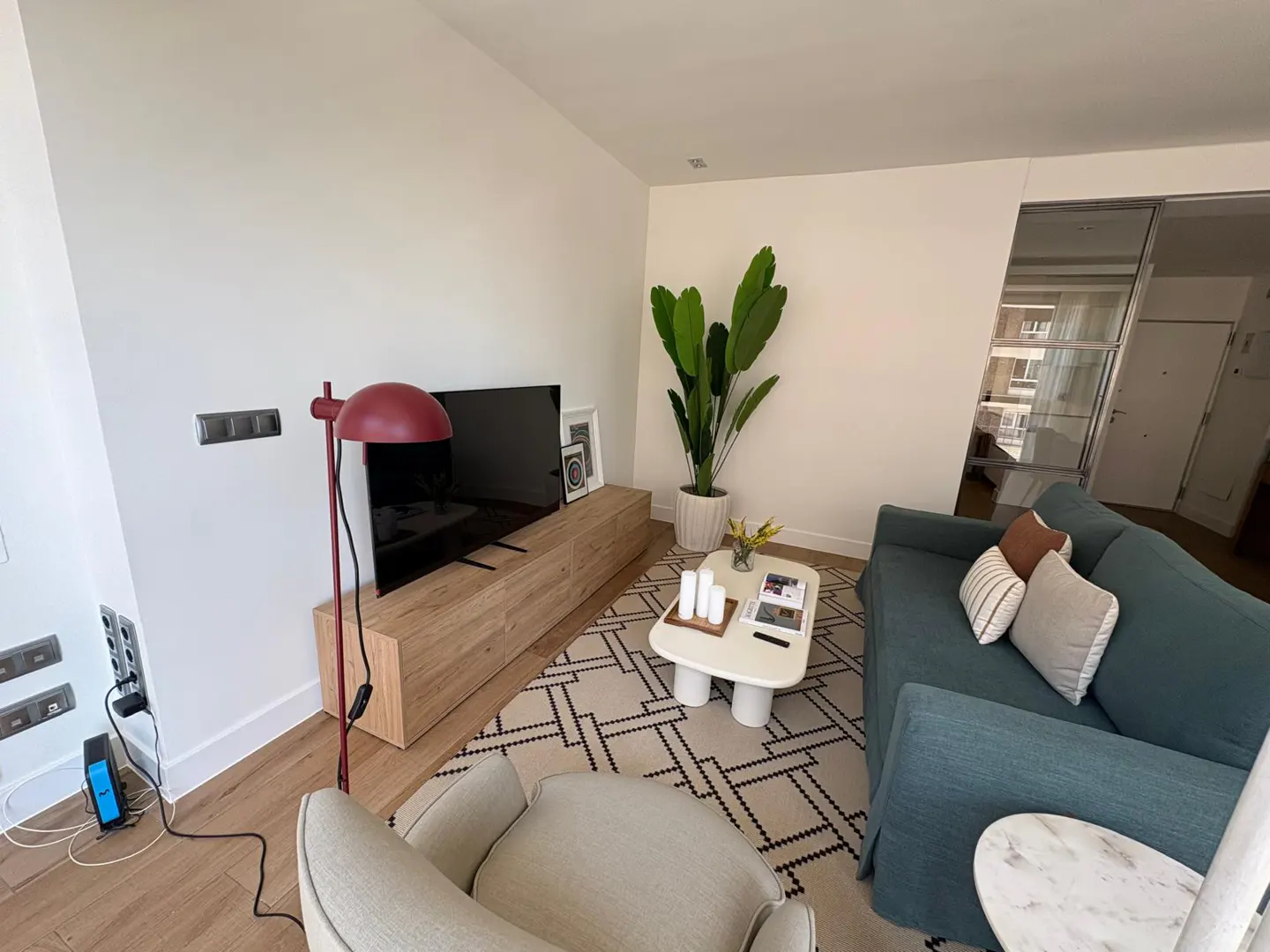Modern living room with a teal sofa, beige armchair, wooden media console, TV, and a geometric patterned area rug.