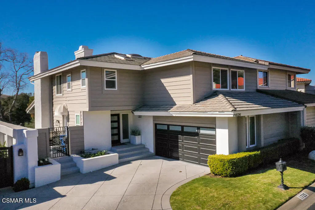 Two-story beige house with a dark garage, white trim, and a concrete driveway under a clear blue sky.