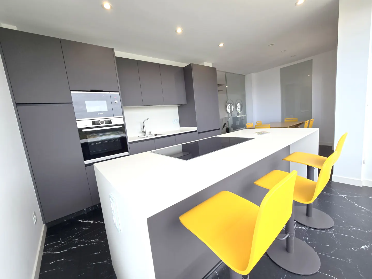 Modern kitchen with gray cabinets, white countertops, and yellow bar stools on a black marble floor.