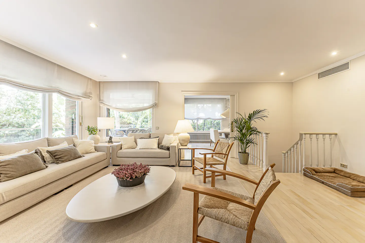 Bright living room with beige sofas, wooden chairs, and a white oval coffee table with a floral centerpiece. Large windows and a staircase are visible.