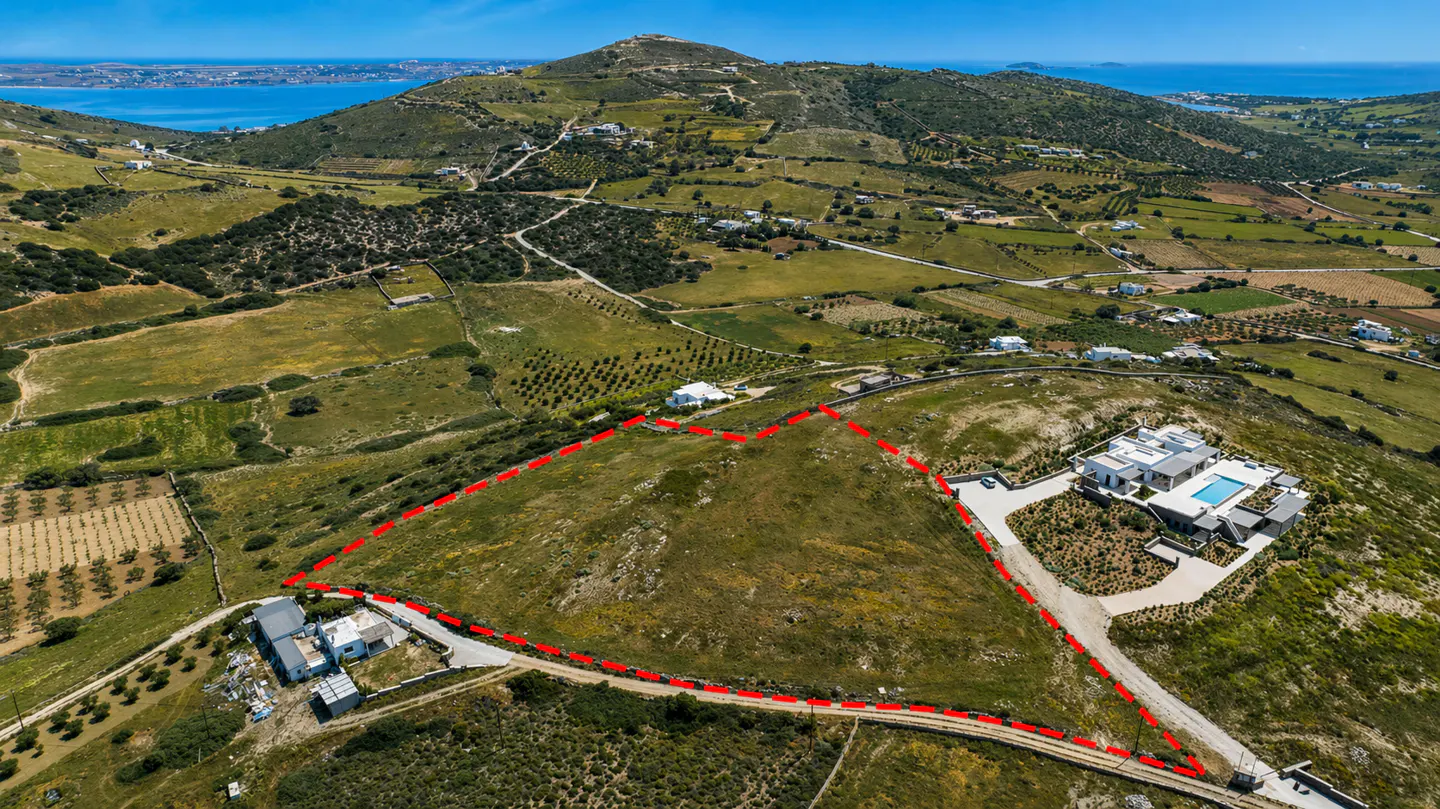 Aerial view of a large plot of land outlined in red, with a modern white house and pool nearby, set against a hilly, green landscape.