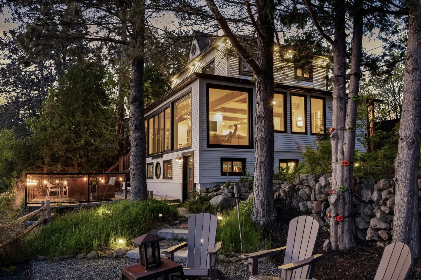 Exterior view of a white house with a sunroom, deck, and Adirondack chairs in the yard.