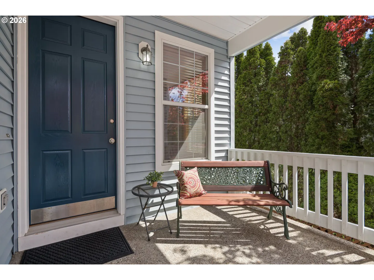 Covered porch with blue siding, a dark blue door, and a brown bench with a patterned pillow. A small table with a plant sits next to the bench.