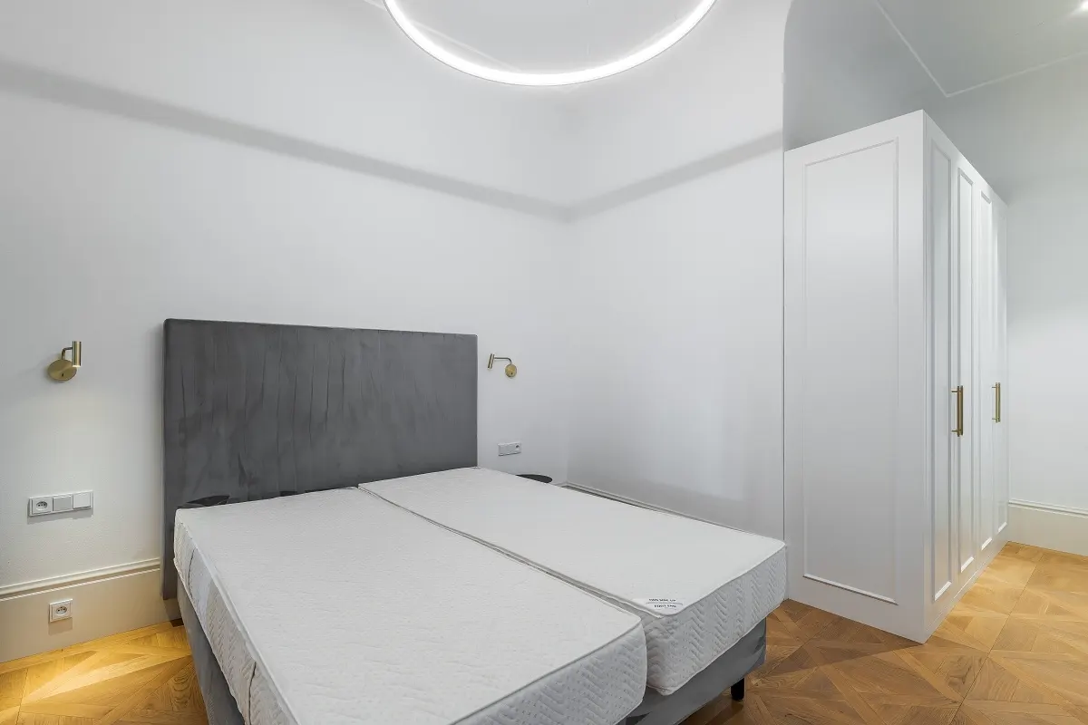 Modern bedroom with two twin mattresses, a grey headboard, white wardrobe, and herringbone wood flooring.
