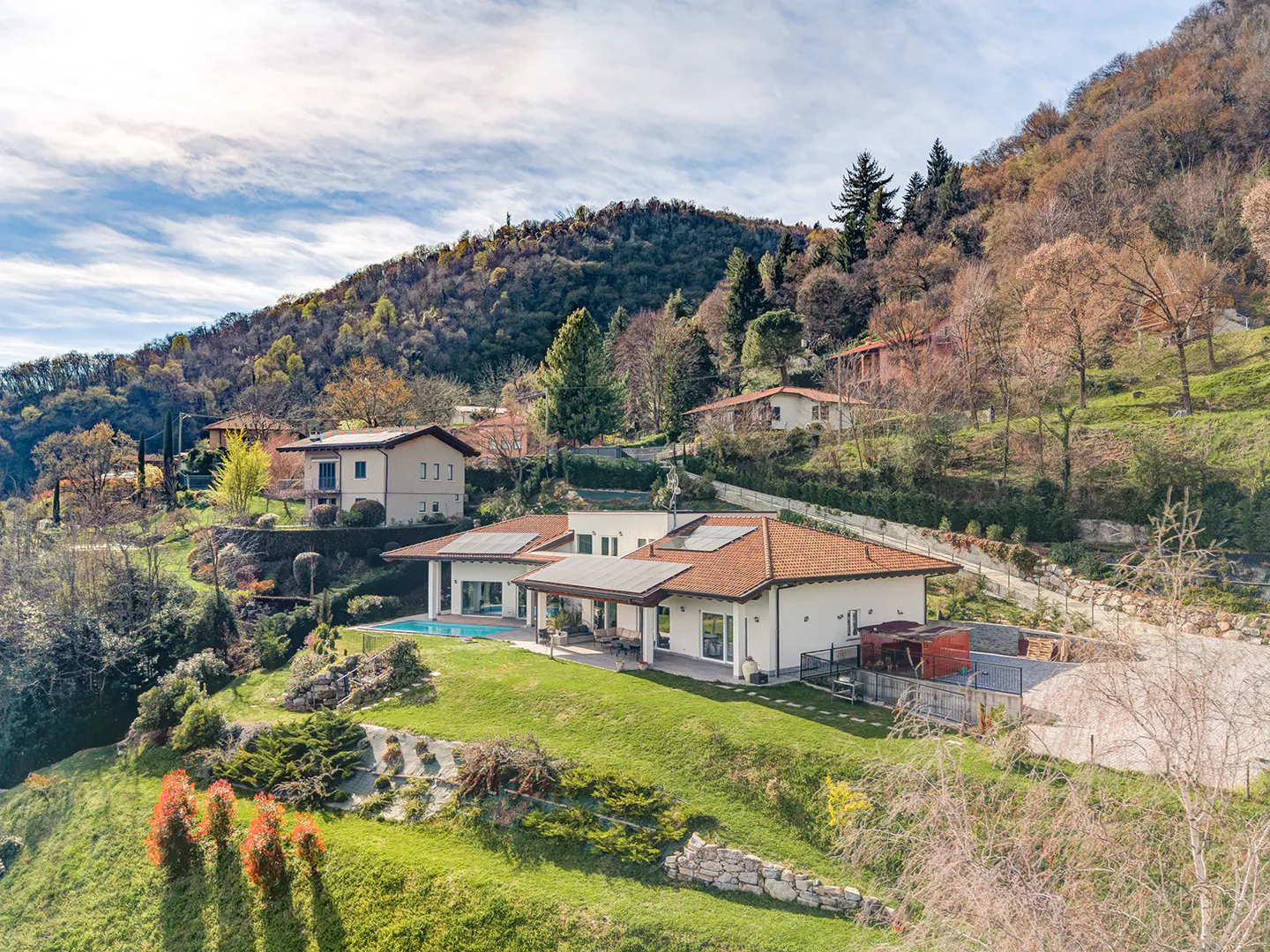 Aerial view of a modern white house with a red tile roof, pool, and green lawn on a hillside.