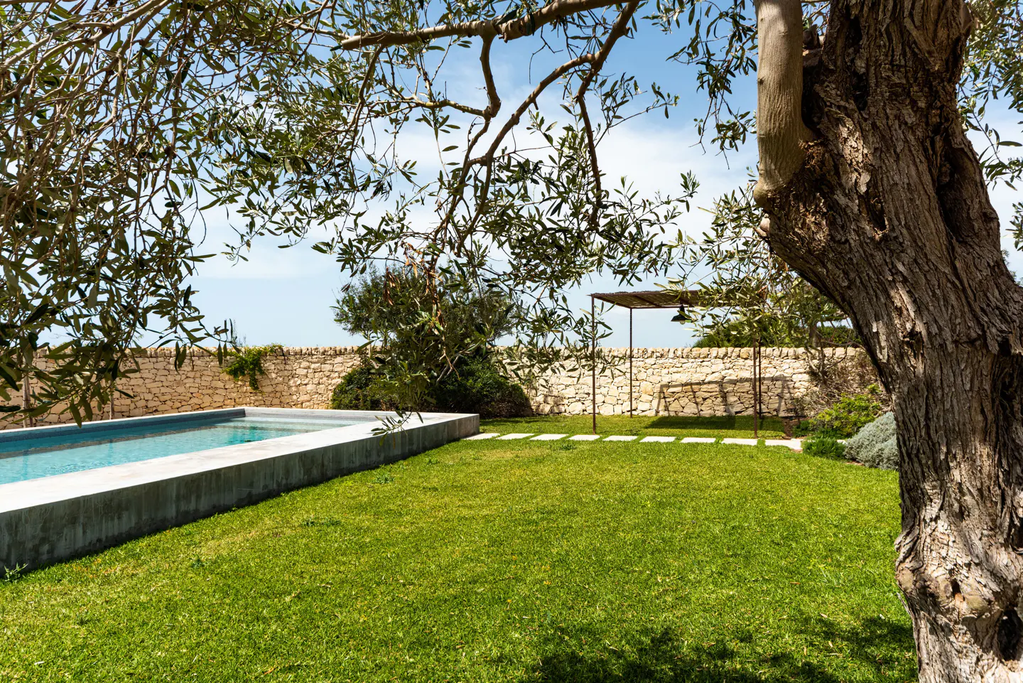 A backyard with a pool, green grass, stone wall, and a metal pergola under a blue sky.