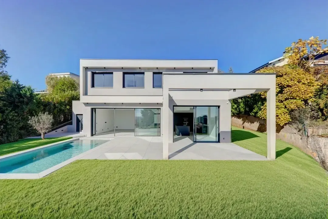 Modern two-story house with a pool. The house is white with black framed windows and sliding glass doors. Green lawn and blue sky.