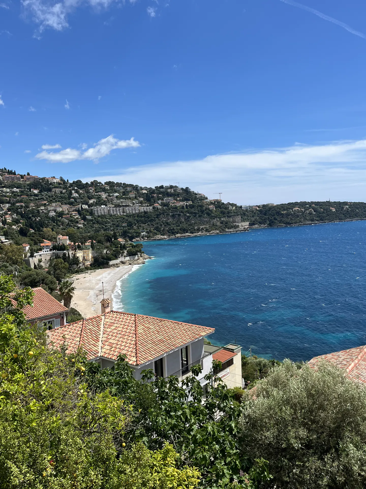 Coastal view of Villefranche-sur-Mer, France, with turquoise water, a beach, and hillside homes under a blue sky.