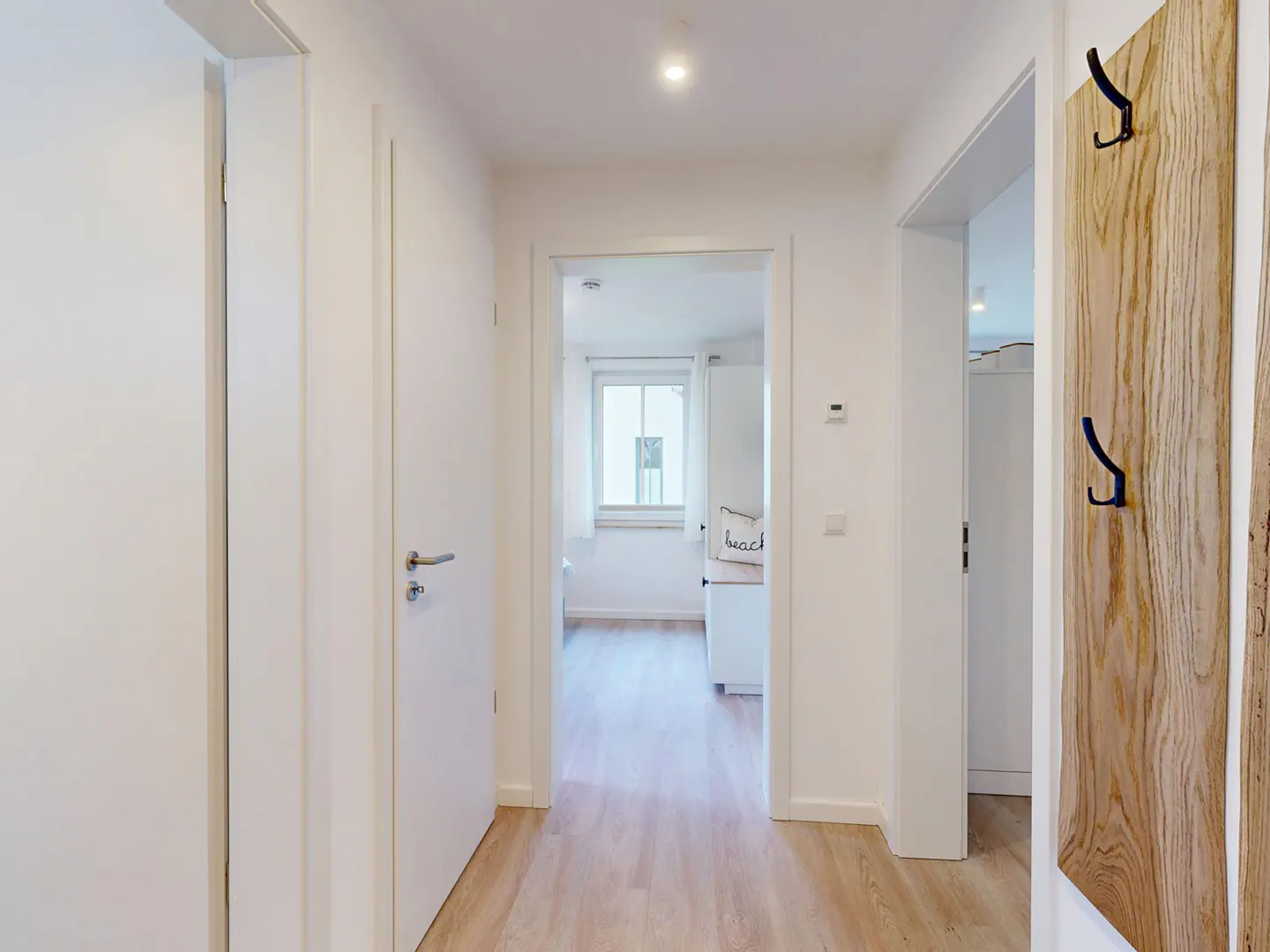 Bright hallway with light wood floors, white walls, and doors. A wood coat rack with black hooks is on the right. A room is visible through a doorway.