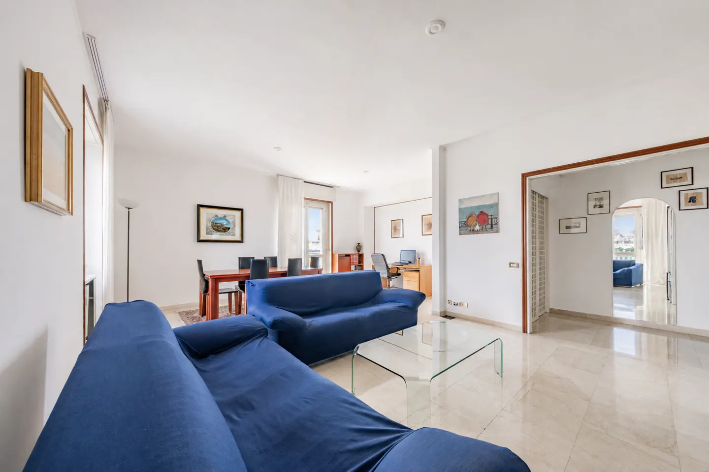 Bright living room with blue sofas, a glass coffee table, and marble floors. A dining table and office area are visible in the background.