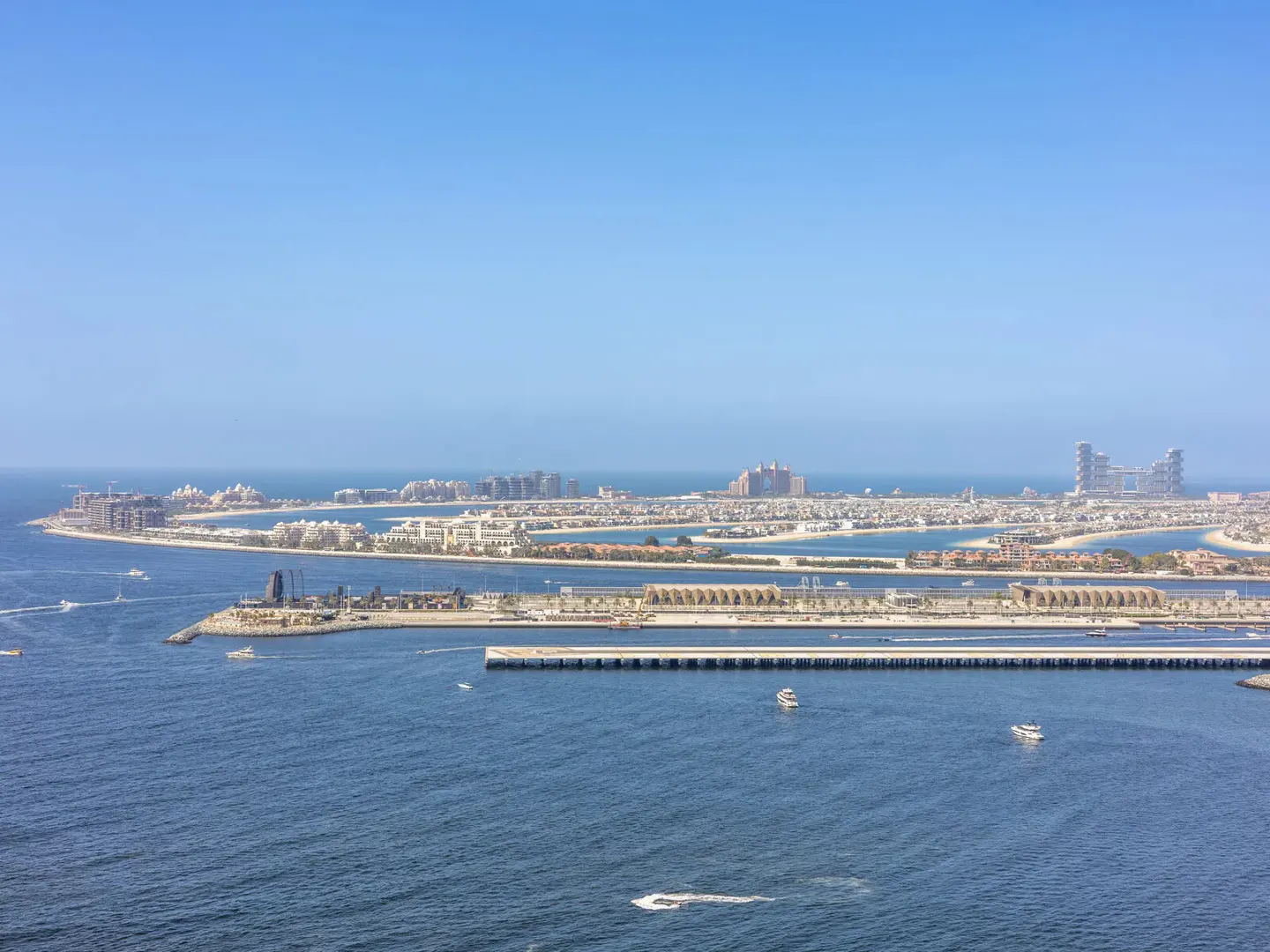 Aerial view of the Palm Jumeirah in Dubai, featuring luxury resorts, a long pier, and boats on the blue sea under clear sky.