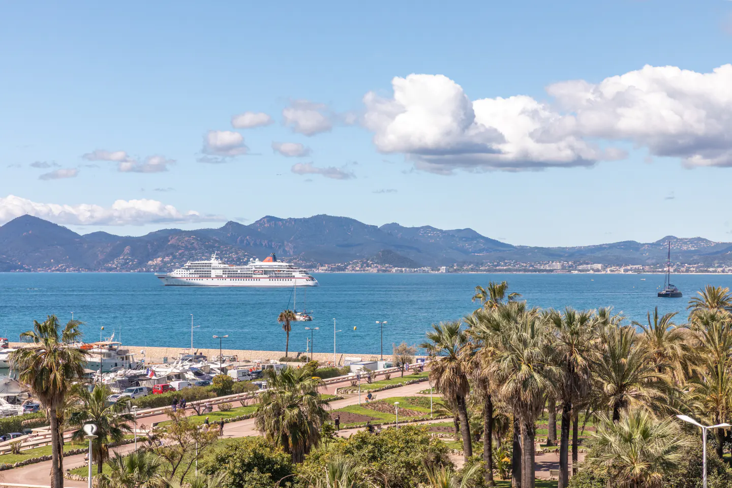 Scenic view of Cannes, France, featuring a cruise ship on blue water, palm trees, and mountains under a partly cloudy sky.