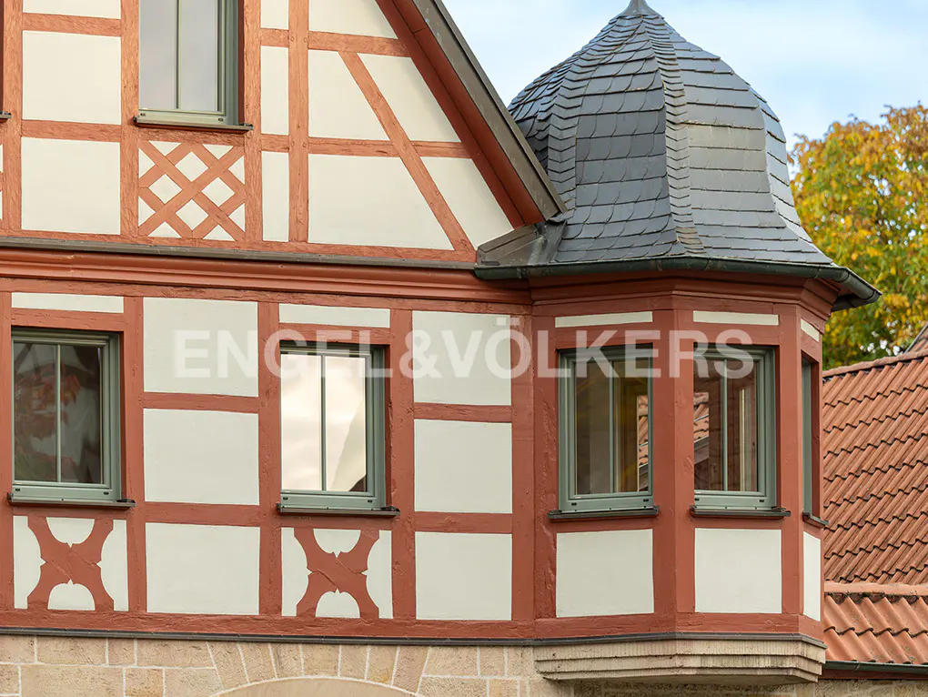 Close-up of a half-timbered house with white panels and red-brown beams, featuring a turret with a gray slate roof. Windows have green frames.