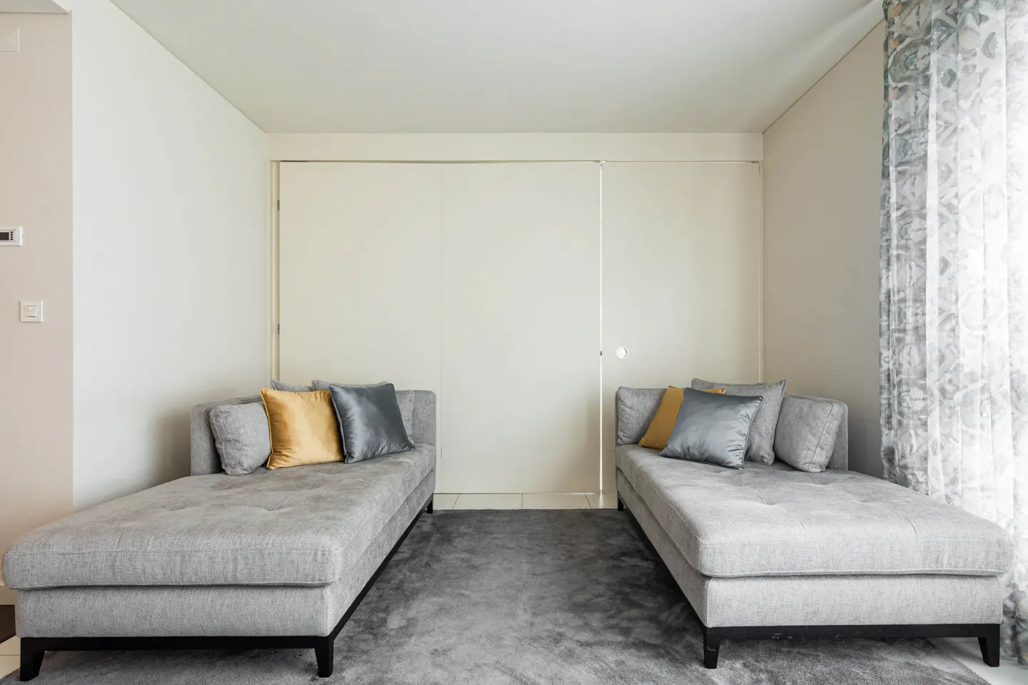 A bright room with two gray chaise lounges facing each other on a gray rug. Each has gold and gray pillows. White walls and patterned curtains.