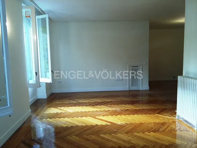 An empty room with herringbone wood floors, white walls, and open windows. The Engel & Völkers logo is visible on the wall.