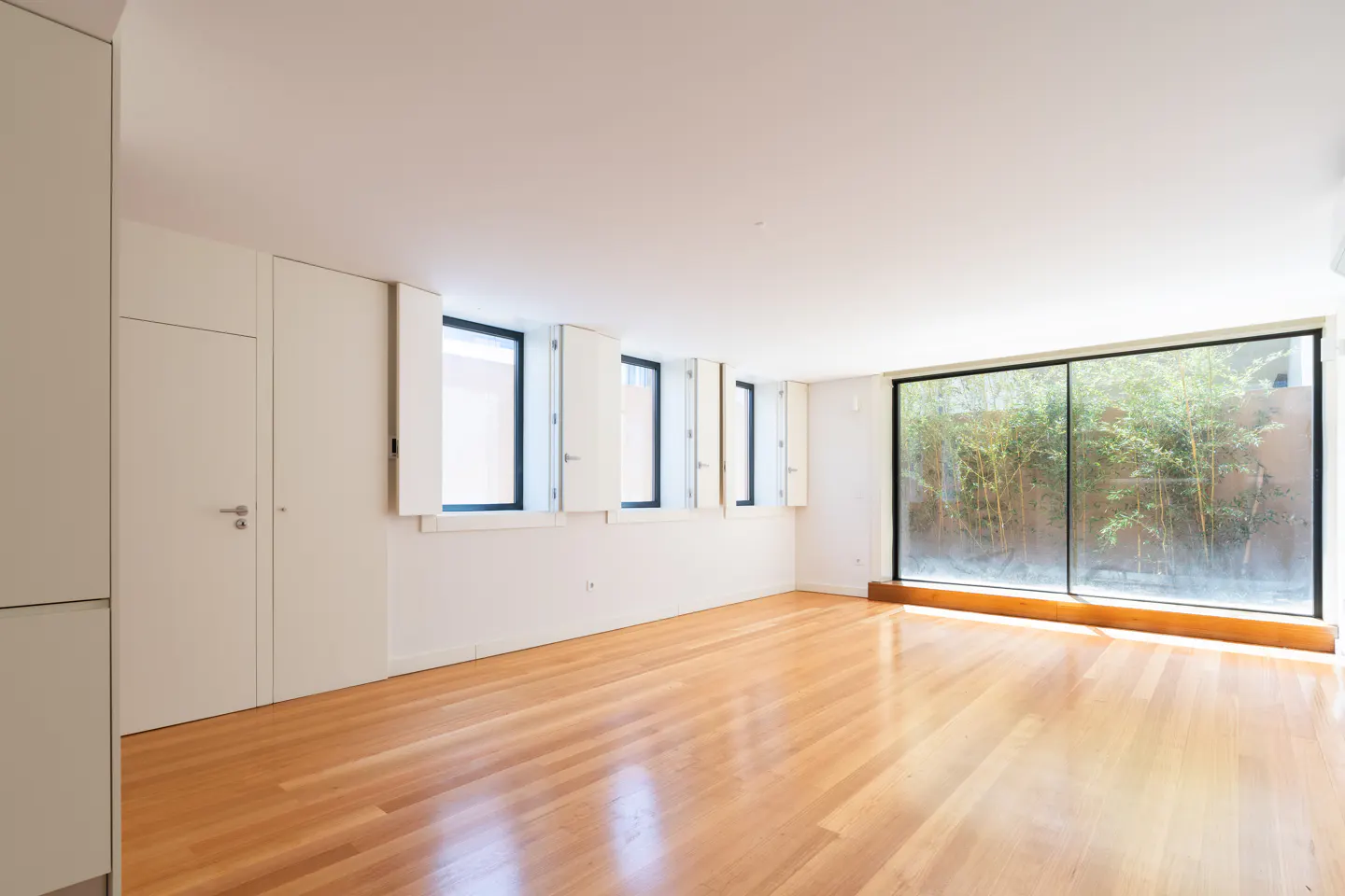 Bright, empty room with wood floors, white walls, and a large window showing greenery outside. Four smaller windows with white shutters line the wall.