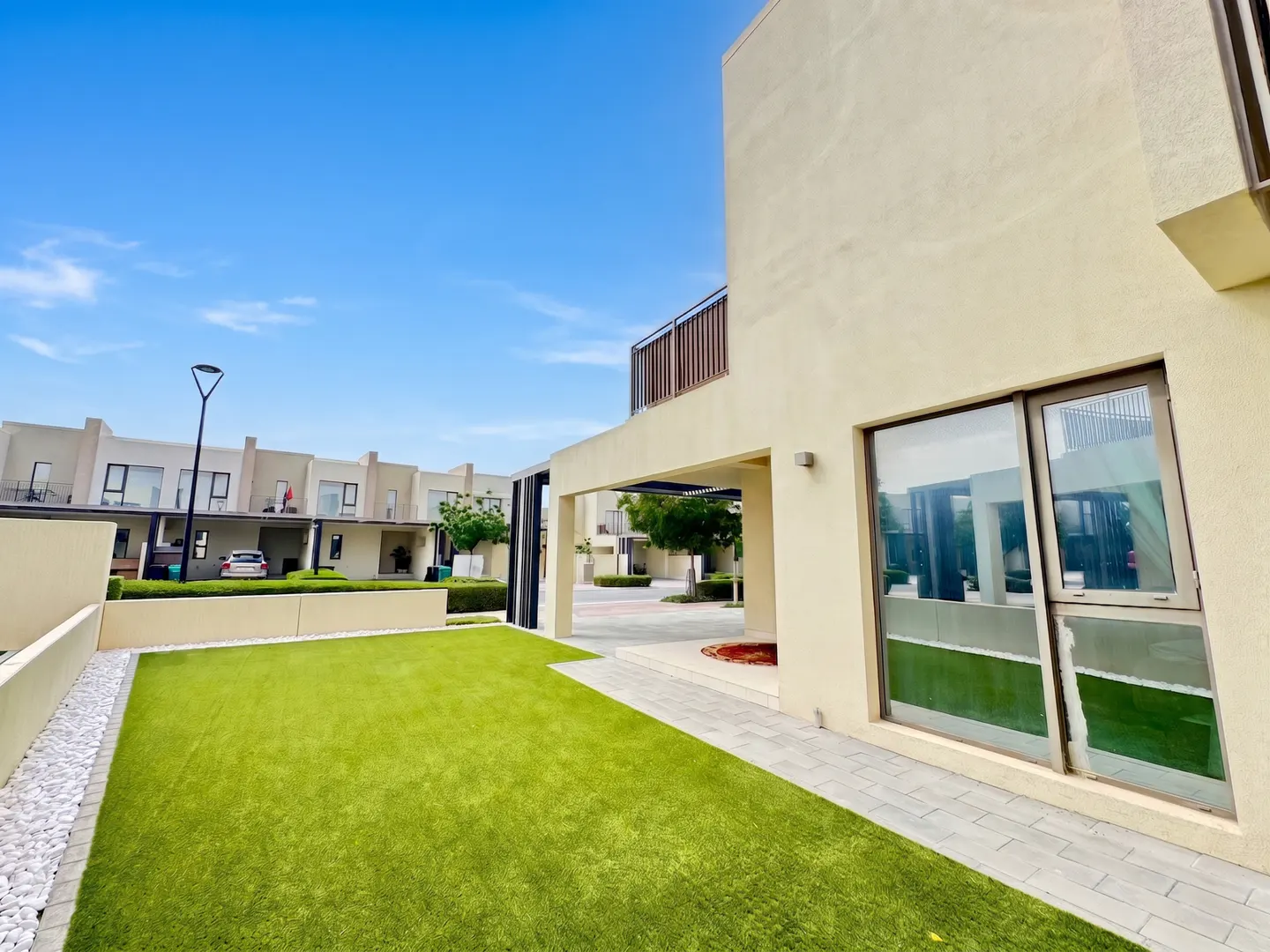 Exterior view of a modern, beige townhouse with a green lawn and blue sky. A large window reflects the lawn and nearby buildings.