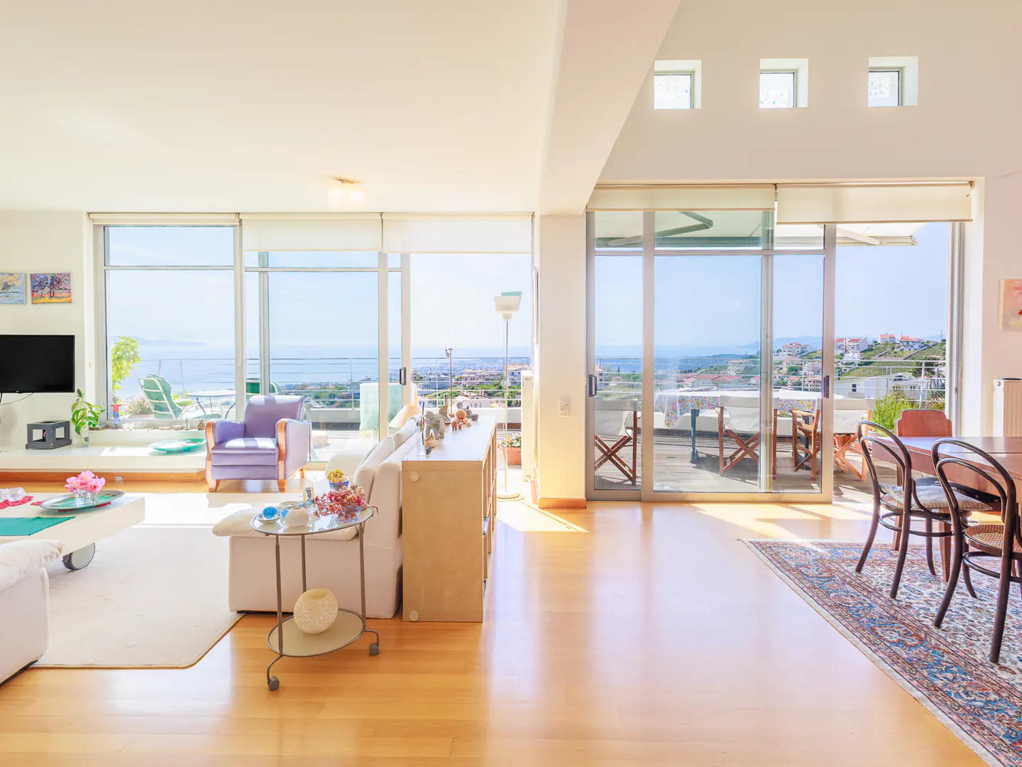 Bright, open living room with hardwood floors, white walls, and large windows overlooking the ocean. Furniture includes a white sofa, purple chair, and dining table.