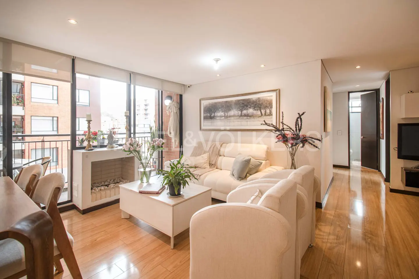 Bright living room with hardwood floors, white sofas, a coffee table, and a faux fireplace with a balcony view.