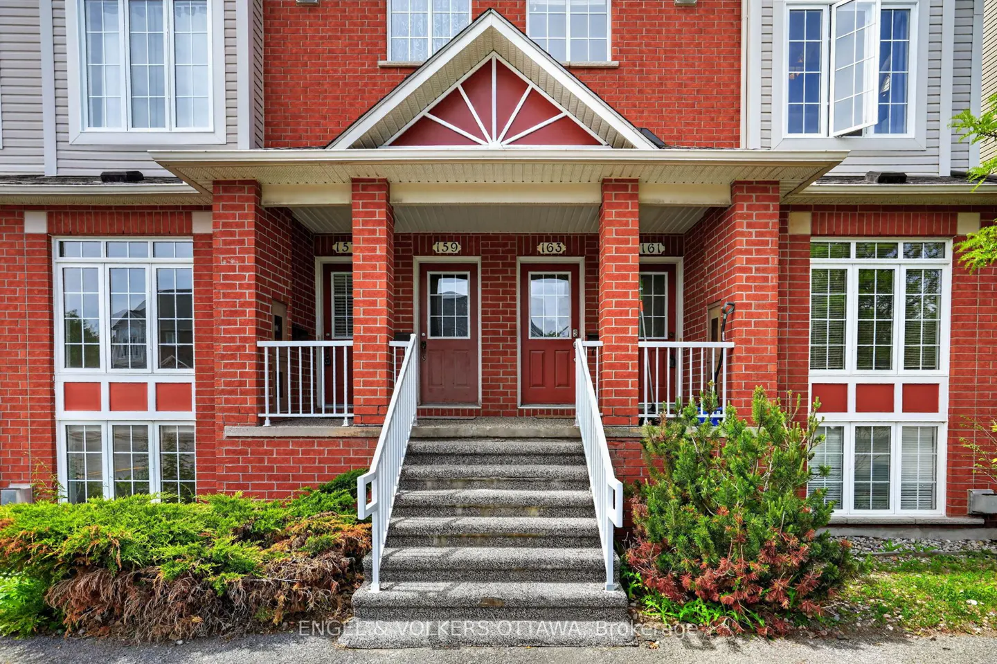 Front view of a red brick townhouse with two entrance doors, concrete steps, white railings, and green landscaping.