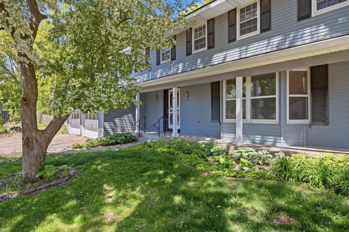 Two-story blue house with black shutters, a covered porch, and a green lawn with a flowering tree.