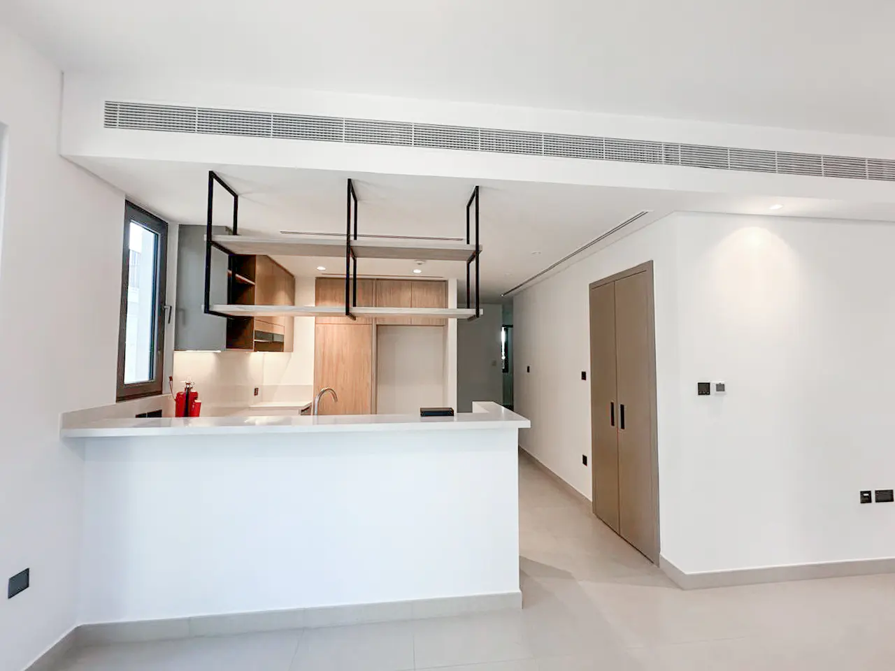 Bright, modern kitchen with white walls, light wood cabinets, and a white countertop island. Black metal shelving hangs above the island. A closed, light brown door is on the right.
