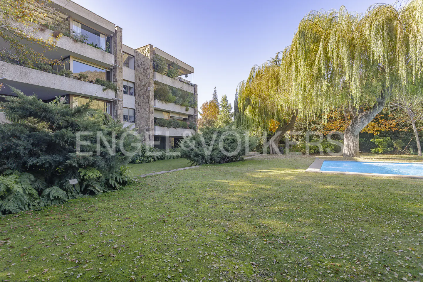 Apartment building with stone accents, balconies with greenery, lawn, pool, and weeping willow tree.