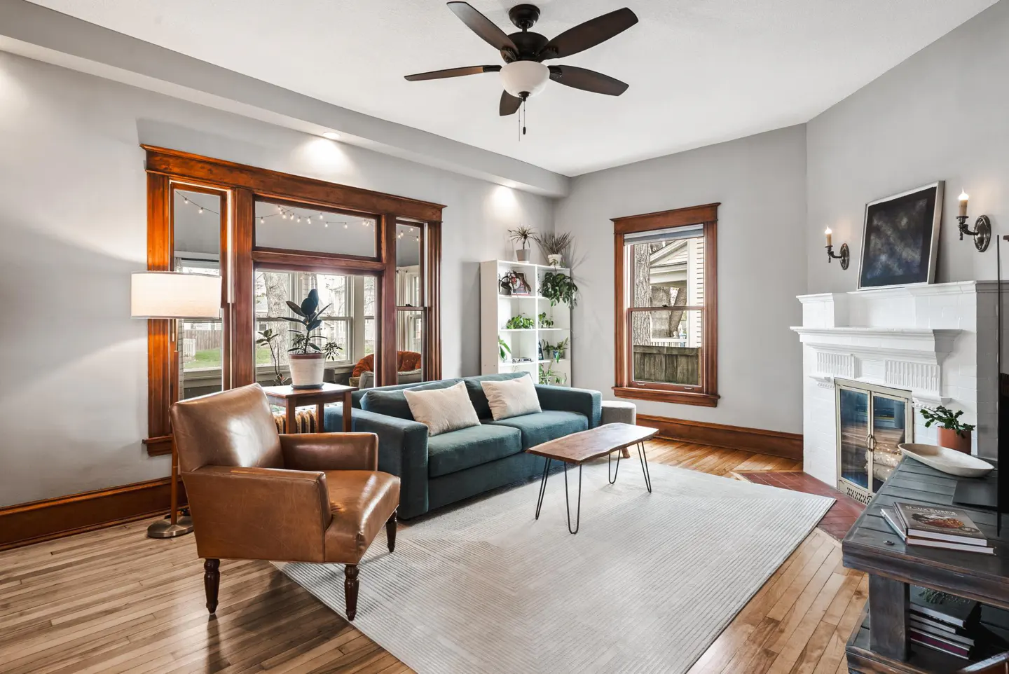 Living room with hardwood floors, a teal sofa, a brown leather chair, and a white fireplace. A ceiling fan hangs above.