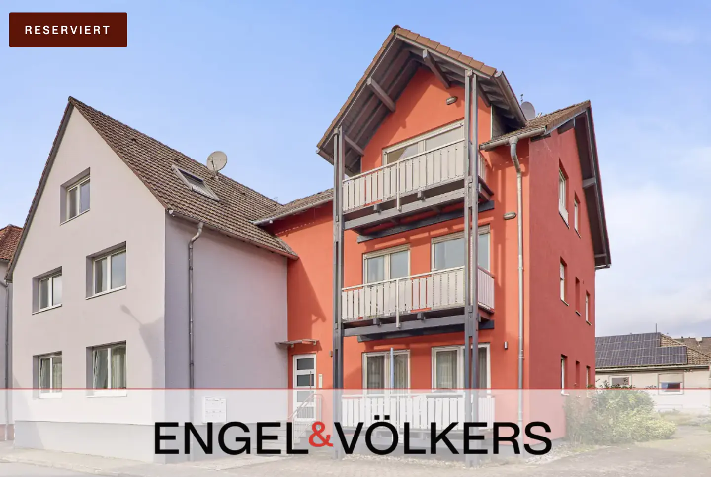 Exterior view of a two-story house with a red facade and balconies, connected to a gray house under a blue sky.