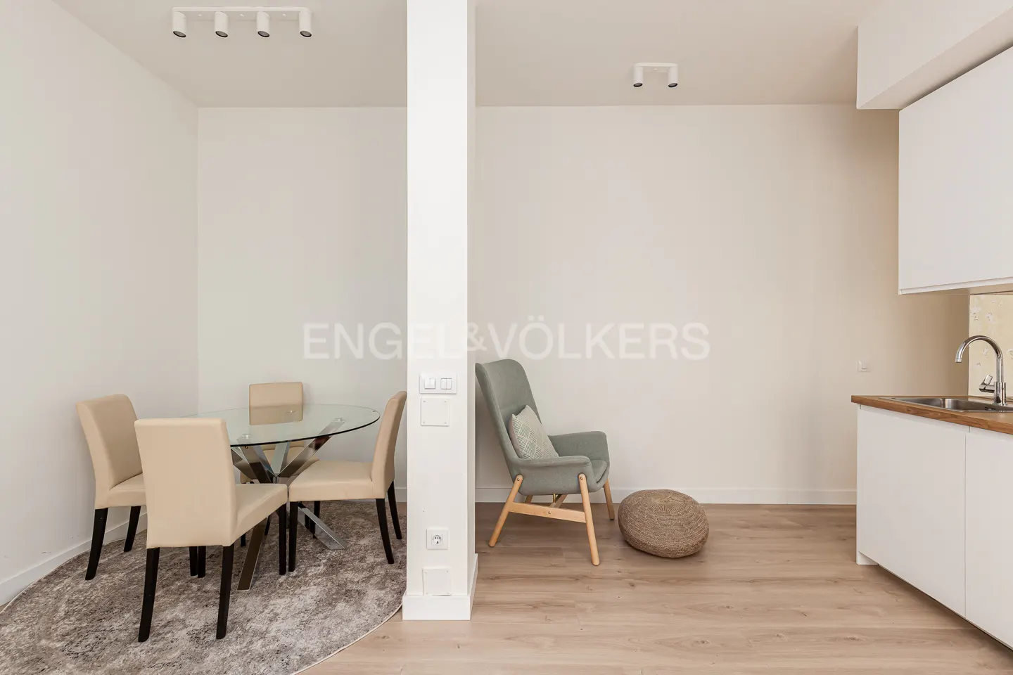 Bright apartment interior with a glass dining table, beige chairs, a gray armchair, and a white kitchen.
