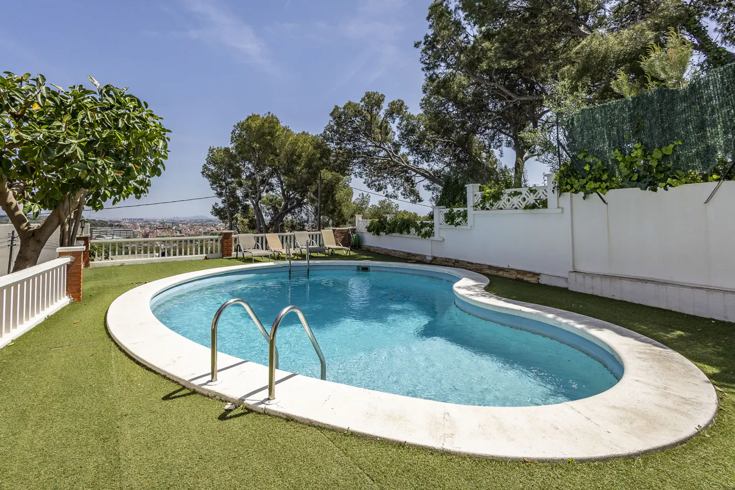 A kidney-shaped pool with turquoise water, surrounded by green turf, sits in a backyard with trees and a white fence.