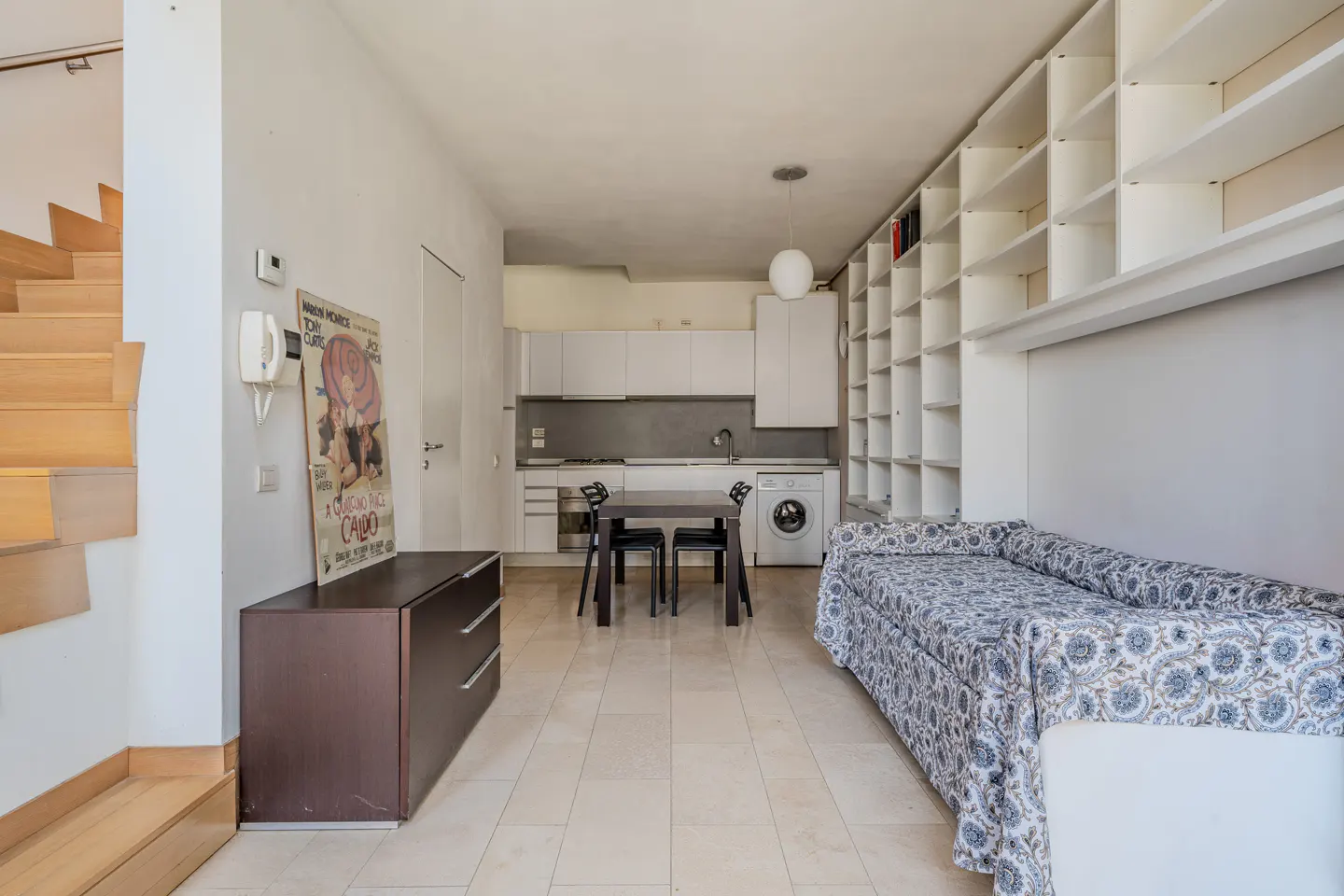 A bright studio apartment with a kitchen, dining table, sofa bed, and a large white bookshelf. A wooden staircase is on the left.
