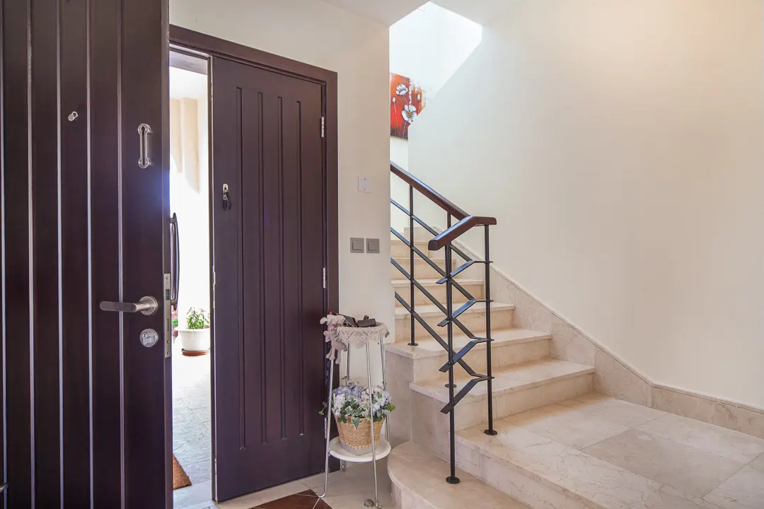 Open brown front door reveals a foyer with marble stairs, a black metal railing, and a small flower stand.