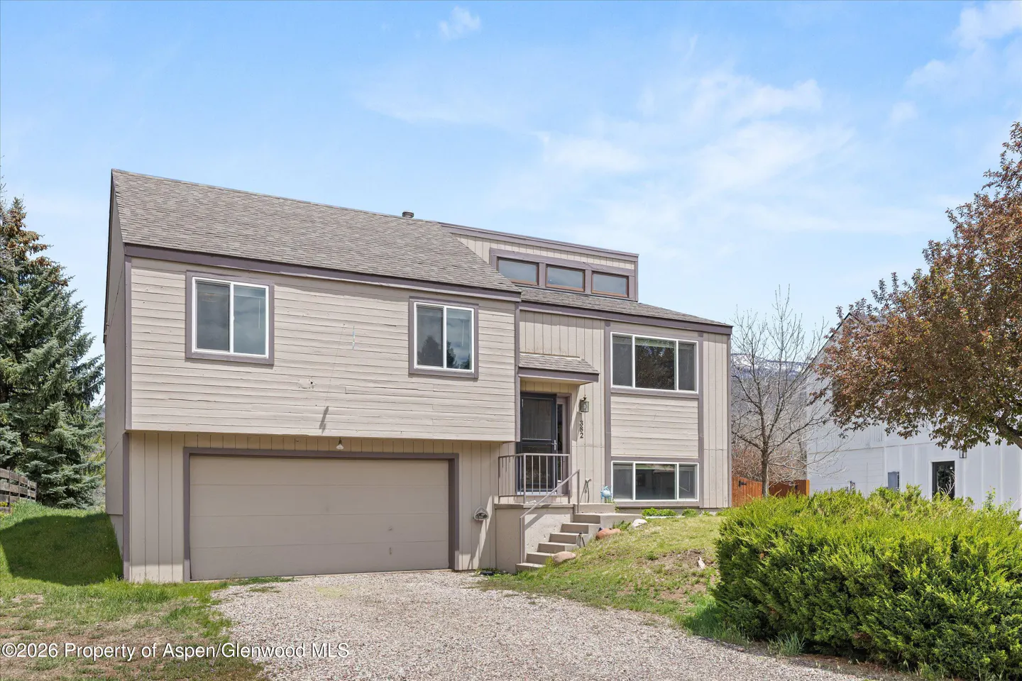 Two-story beige house with a gray roof, garage, and gravel driveway on a sunny day.