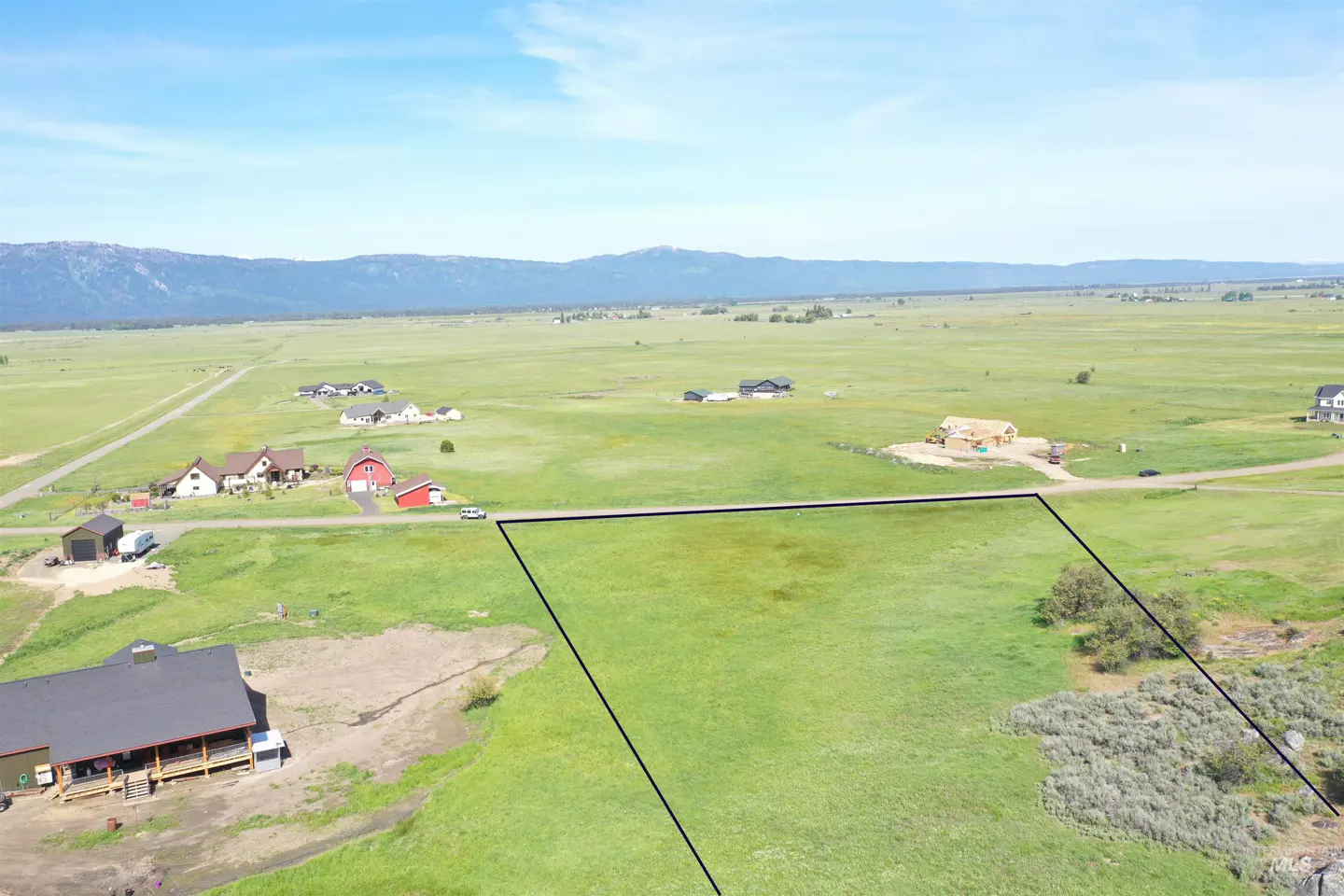 Aerial view of a grassy lot outlined in black, with houses, fields, and mountains in the background under a blue sky.