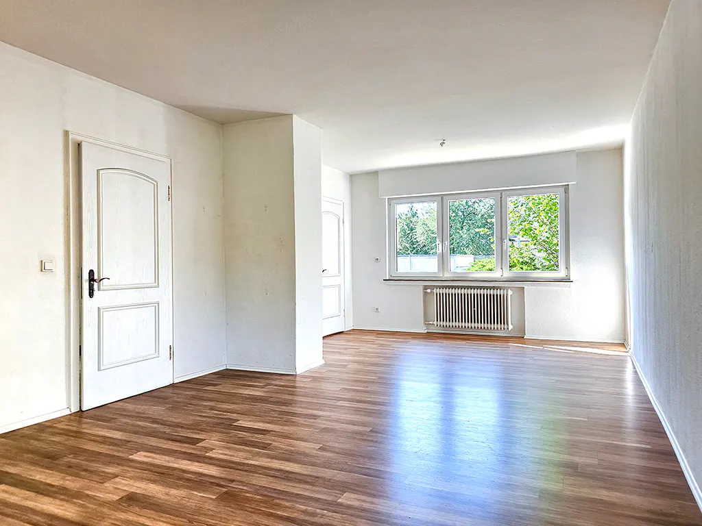 Bright, empty room with wood floors, white walls, and a window showing green trees. A white door is on the left.