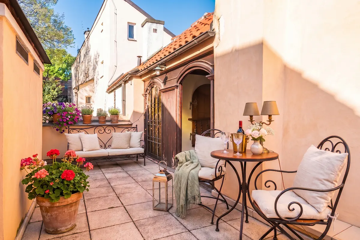 Cozy outdoor patio with wrought iron furniture, a small table set with wine, and a bench with flowers. Peach-colored walls surround the space.