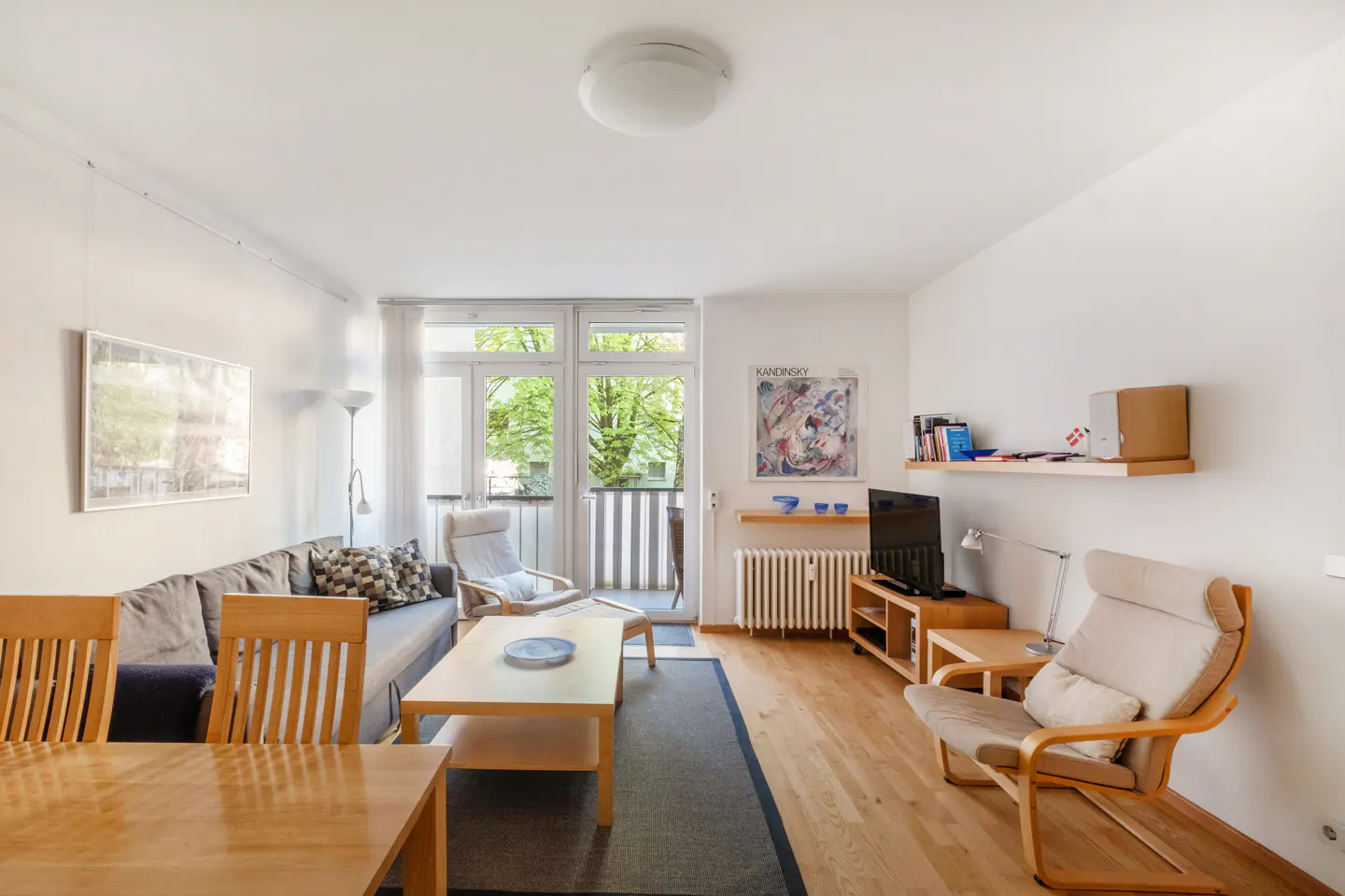 Bright living room with wood floors, gray sofa, chairs, and a balcony with green trees visible outside.