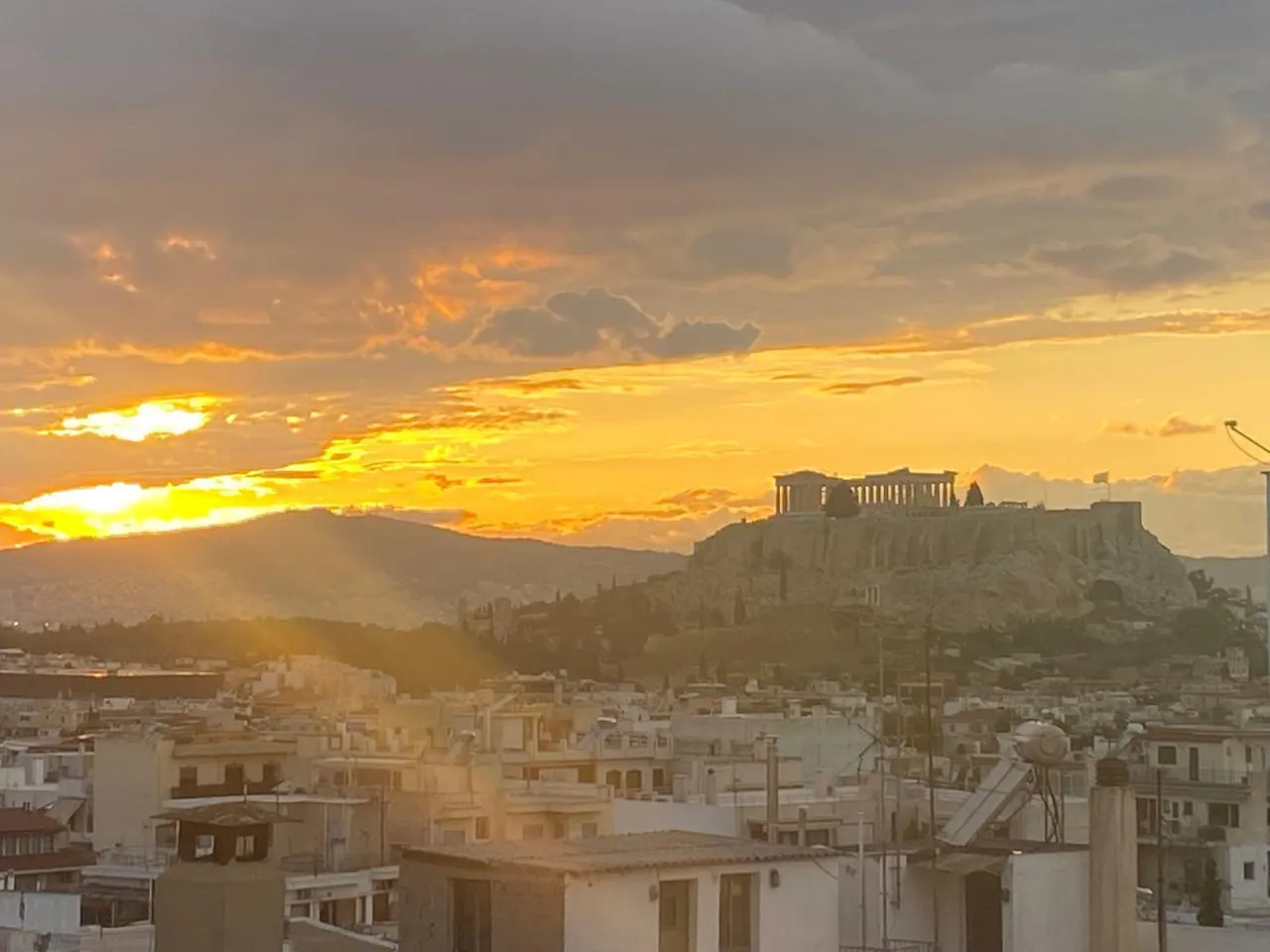Athens cityscape at sunset. The Acropolis sits atop a hill overlooking white buildings under a golden sky with sun rays.