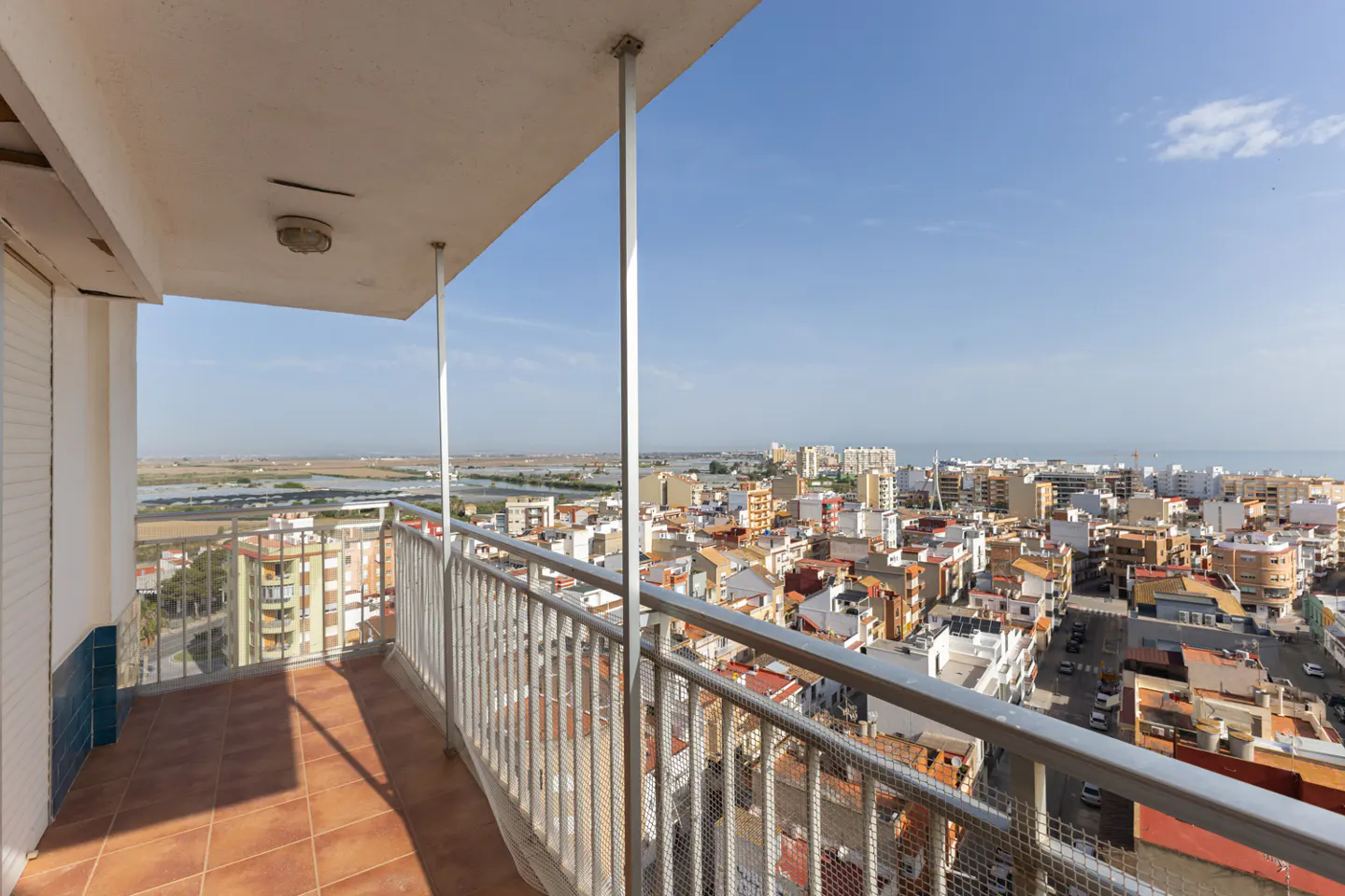 Balcony view overlooking a city with red-tiled roofs, with a blue sky and distant water. The balcony has brown tiled floor and metal railings.