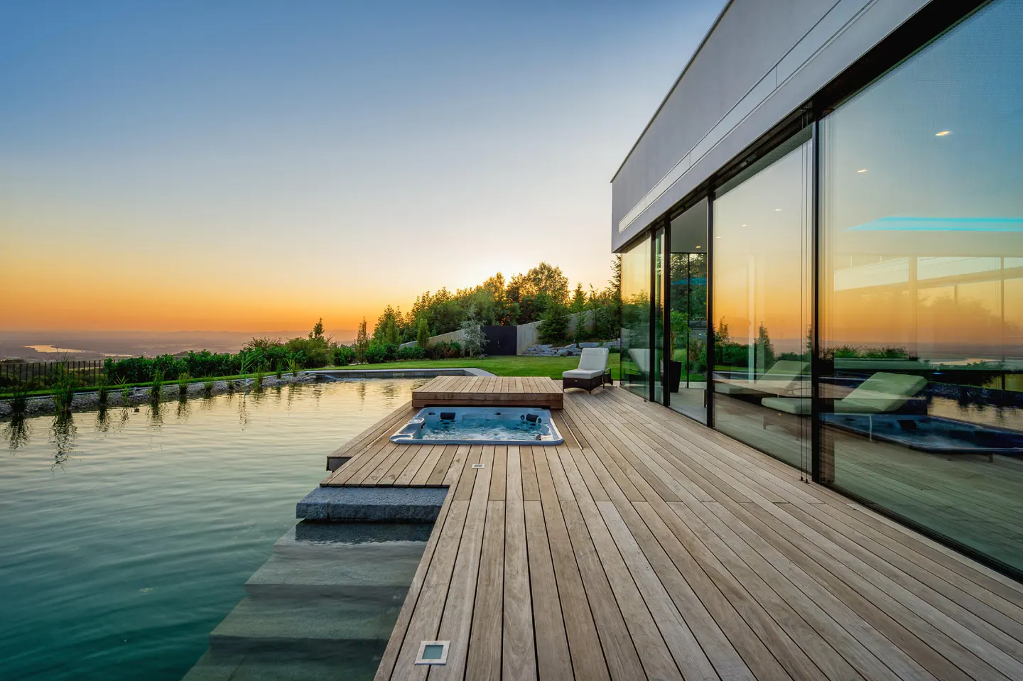 Modern home exterior with a pool, hot tub, and wooden deck at sunset. Large glass doors reflect the orange sky.
