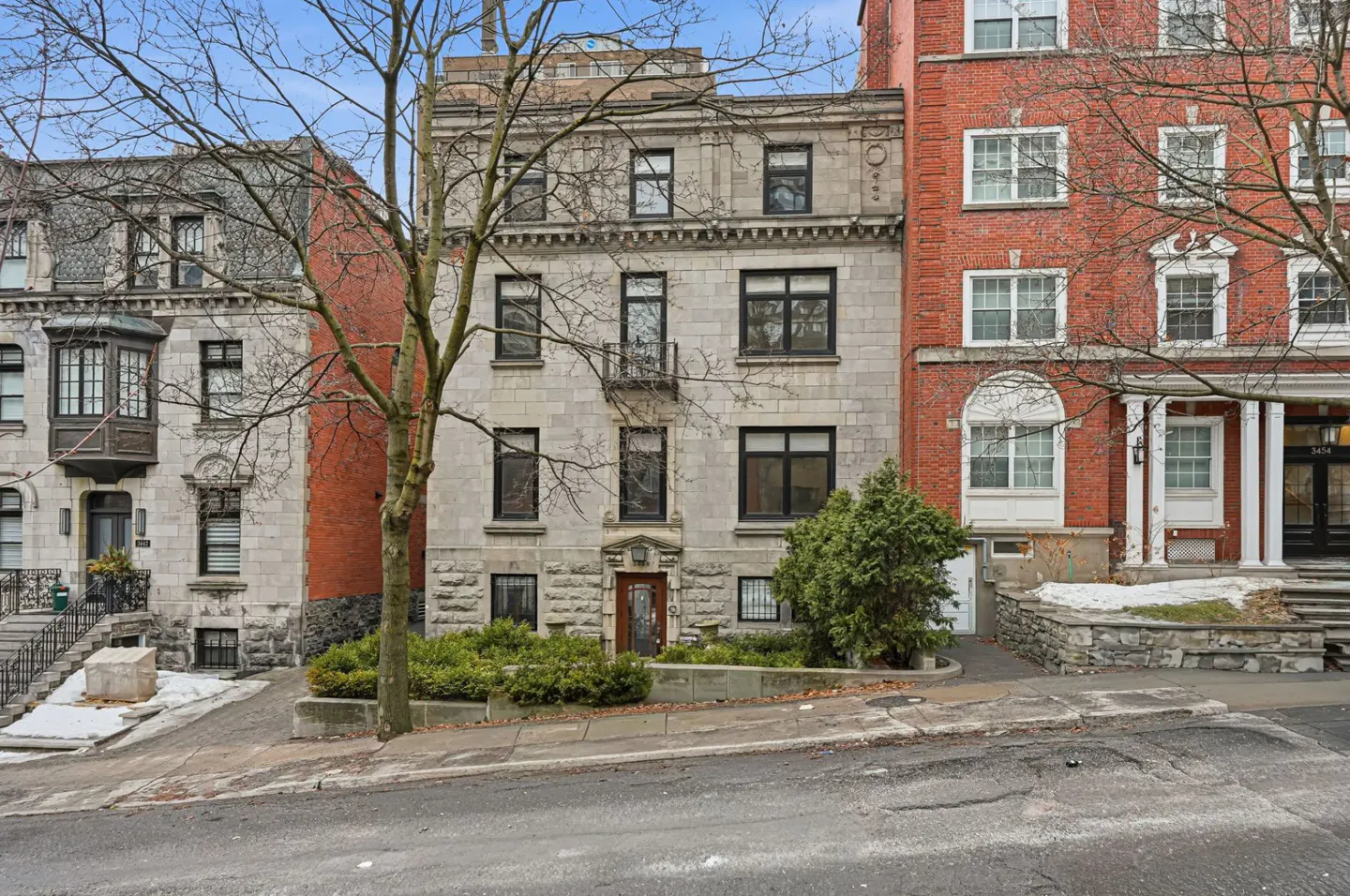 Exterior view of a three-story stone building with black-framed windows, flanked by brick buildings and bare trees.