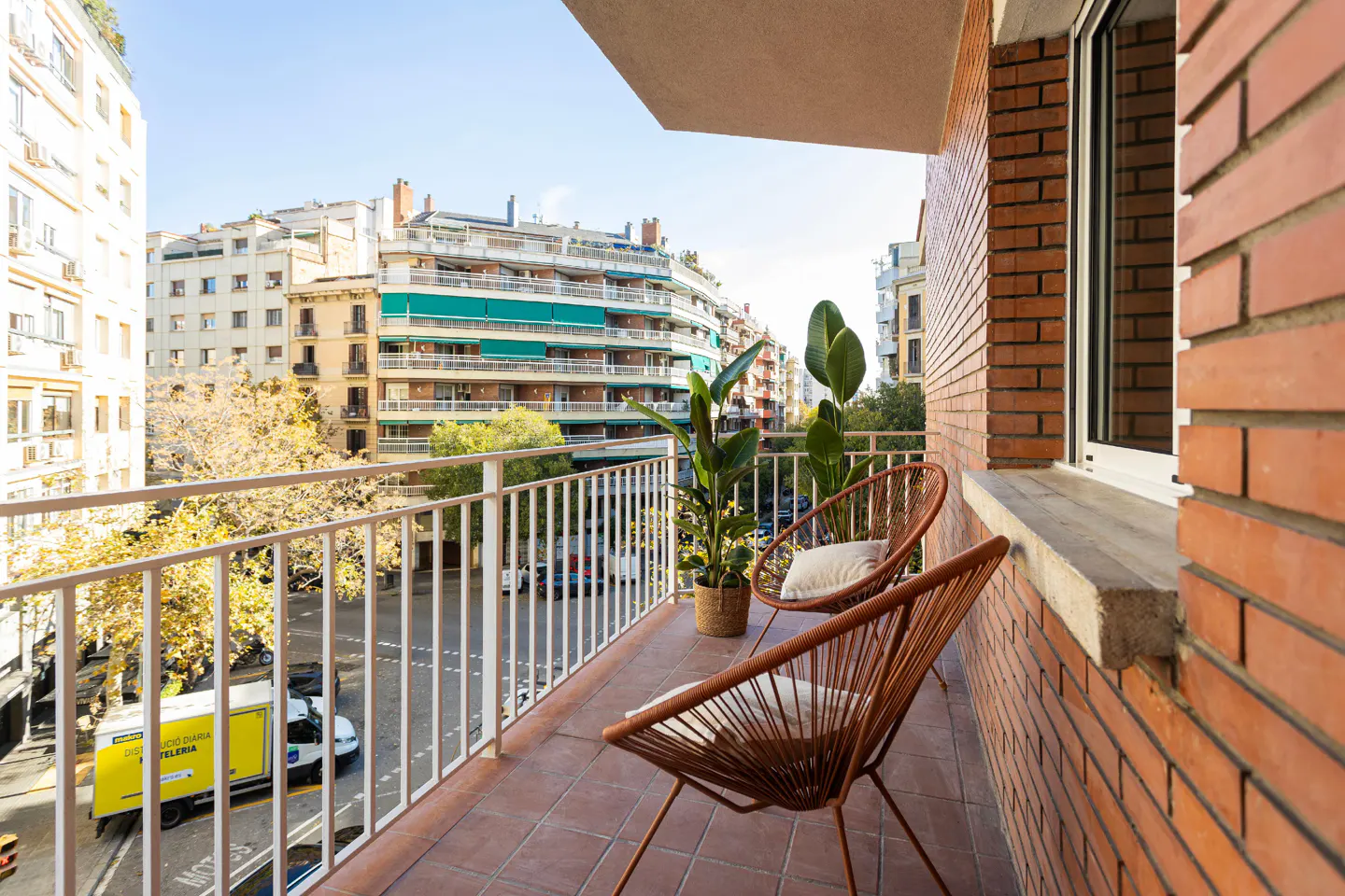 Balcony view with two brown chairs, white cushions, and plants. White railing overlooks a city street with buildings and traffic.