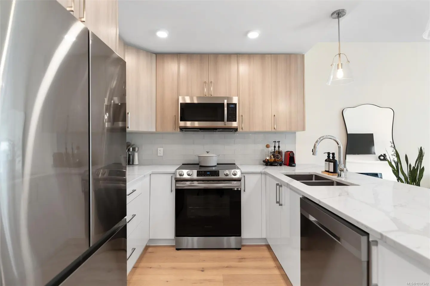 Bright kitchen with stainless steel appliances, light wood cabinets, and white countertops. A mirror and plant are visible in the background.