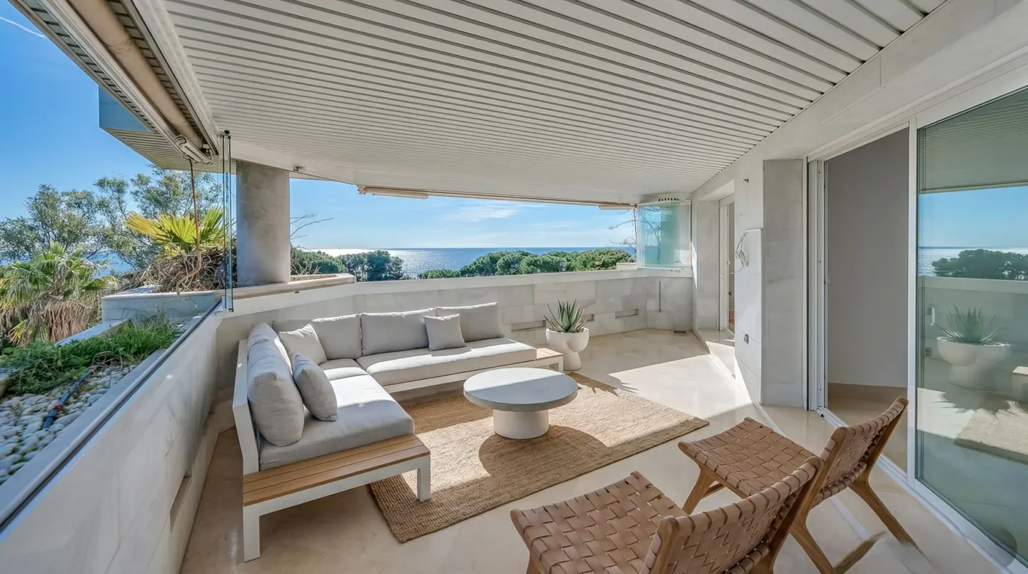 A bright, modern balcony with a gray sectional sofa, round table, woven rug, and two chairs overlooking the ocean.