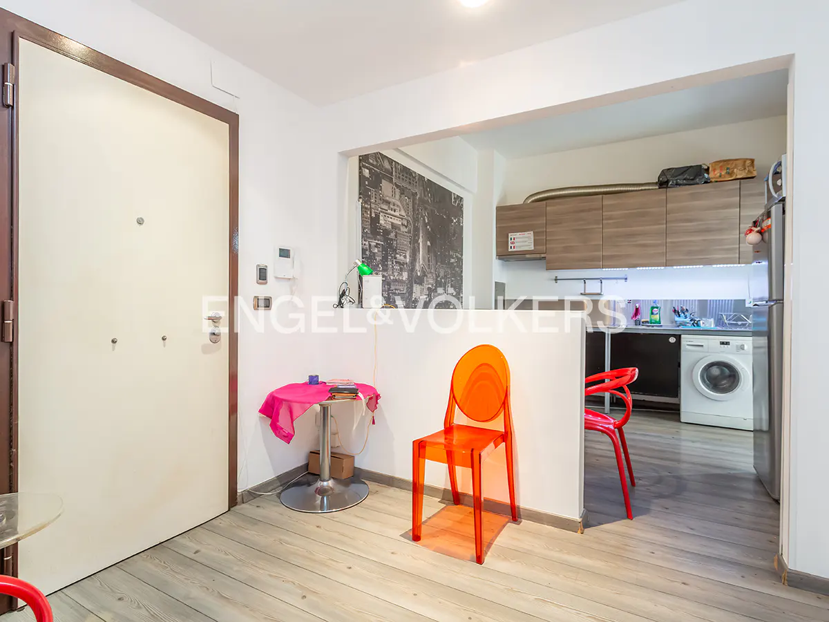 A bright apartment interior with a white door, orange chairs, and a kitchen view. The floor is light wood.